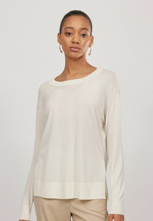 Kvinde iført en løs, langærmet cremefarvet sweater med ribbet kant og manchetter, kombineret med beige bukser, stående mod en ensfarvet baggrund.