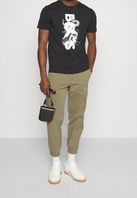 Svart grafisk t-shirt med vitt mönster, olivgröna cargobyxor, vita sneakers med texturerad yta och en liten svart väska.