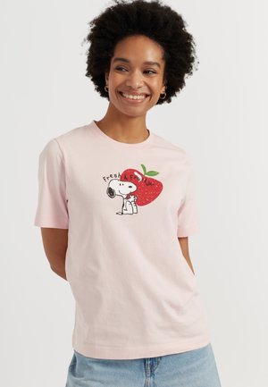 Donna sorridente con capelli corti e ricci che indossa una T-shirt rosa chiaro con Snoopy seduto davanti a una grande fragola rossa e la scritta "Fresh & Fruity".