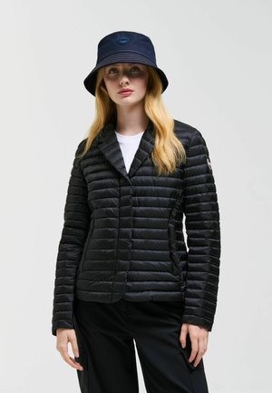 Giovane donna con lunghi capelli biondi indossa una giacca trapuntata nera, cappello a secchiello blu scuro, camicia bianca e pantaloni neri su sfondo uniforme.