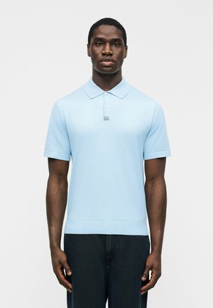 Mand stående vendt fremad iført en lyseblå poloshirt med korte ærmer og mørke bukser mod en ensfarvet hvid baggrund.