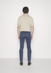 Camiseta beige de manga corta combinada con jeans de denim azul ajustados y zapatos negros, vista desde atrás sobre un fondo blanco.
