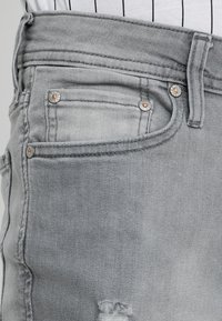 Acercamiento del área del bolsillo de unos jeans de mezclilla gris con costuras visibles, remaches, pasador de cinturón y un desgarro deshilachado cerca del borde inferior.