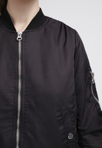 Svart bomberjacka med en slät tygtextur, som har en silverfärgad framdragkedja, ribbad krage och en sidoficka med tryckknapp.
