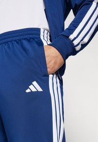 Mornarska atletska trenirka z belimi črtami in Adidas logotipom. Ima elastičen pas in stranske žepke. Gladek, lahek material.
