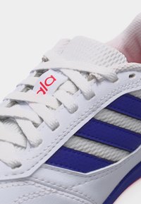 Κοντινό πλάνο λευκού αθλητικού παπουτσιού με μπλε ρίγες, λευκά κορδόνια και ετικέτα στη γλώσσα με κόκκινο κείμενο.