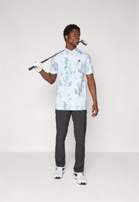 Golf shirt with a light blue and green abstract pattern, short sleeves, and a collar; paired with dark trousers and white athletic shoes.