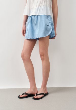 Kobieta ubrana w jasnoniebieskie, pionowo paskowane szorty, czarne klapki i biały bezrękawnik, stojąca na jednolitym tle.