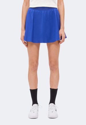 Person steht und trägt einen leuchtend blauen sportlichen Rock, schwarze knielange Socken, weiße Turnschuhe und ein weißes Oberteil, gezeigt von der Taille bis zu den Füßen.