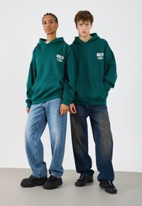 Grüne Kapuzenpullover mit Fronttaschen und weißen Logos, kombiniert mit hellblauen und dunkelblauen weiten Jeans und klobigen schwarzen Schuhen.