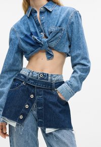 Denim krekls ar sasietu jostas daļu, kas izstrādāts gaismas zilā un tumši zilā krāsu shēmā. To pavada daudzslāņu denim svārki ar pogu akcentiem.