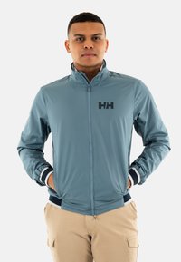 Giacca di jeans leggera di colore azzurro, con zip e colletto alto, dotata di un logo nero sul petto e dettagli a righe sui polsini.