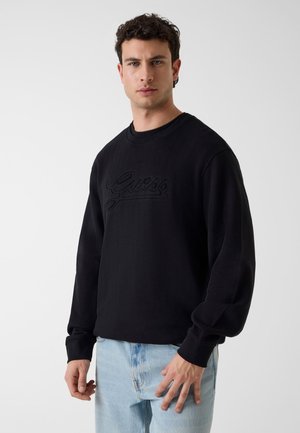 Młody mężczyzna ubrany w czarną bluzę Guess i jasnoniebieskie dżinsy, stojący na jednolitym białym tle.