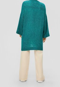 Türkiser übergroßer Strickpullover mit fallenden Schultern, gerippten Bündchen und einem hohen Kragen, kombiniert mit beigen weit geschnittenen Hosen und weißen Sneakern.