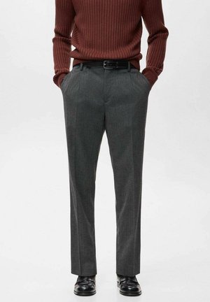 Person trägt braunen Rippstrickpullover, graue maßgeschneiderte Hose mit schwarzem Gürtel und schwarze Lederschuhe, steht mit den Händen in den Taschen.