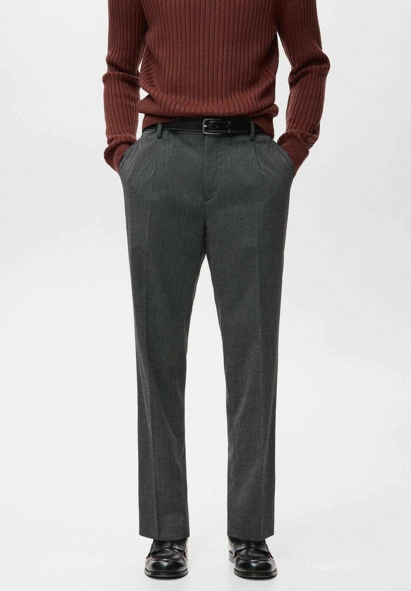 Personne portant un pull en maille côtelée marron, un pantalon gris sur mesure avec une ceinture noire et des chaussures en cuir noires, debout, les mains dans les poches.