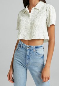 Cropped white shirt with textured fabric, short sleeves, and button closure. Paired with blue high-waisted jeans featuring side pockets.