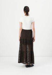 Lang brun blonde linstoffkjole med lagdelt design og gjennomsiktig tekstur, kombinert med en tettsittende hvit kortermet topp og svarte sko.