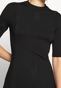 Gros plan sur une personne portant un haut en tricot noir à manches courtes ajusté, avec des motifs subtils en dentelle verticale sur le devant et les manches.