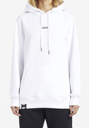 Persoon in een witte oversized hoodie met een voorkussen en een zwarte labeldetail aan de zoom, gecombineerd met zwarte broek.