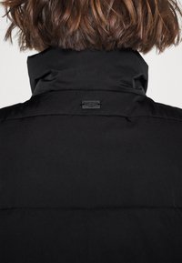 Czarna puchowa kurtka z wysokim kołnierzem i małym, przeszywanym logo na plecach. Matowe wykończenie, gładka faktura oraz widoczne przeszycia.