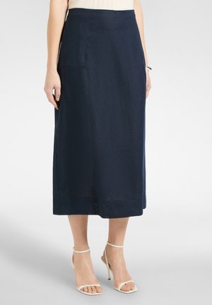 Frau trägt einen marineblauen Midirock kombiniert mit weißen, hochhackigen Sandalen mit Knöchelriemen und steht vor einem schlichten Hintergrund.