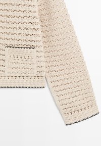 Kremfarvet hæklestrikket langærmet trøje med lille frontal lomme og sort kantbesætning, lagt fladt på hvid baggrund.