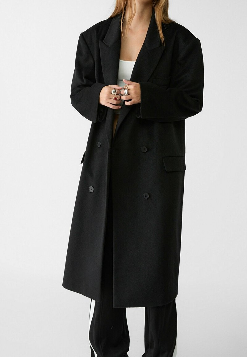 Personne portant un long manteau noir croisé sur un pantalon noir et un haut blanc, montrant des bagues en argent et en or aux deux mains.