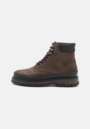 Suede brown ankle boot with black rubber sole. Features metallic eyelets, laces, and padded collar for comfort. Textured surface detail.