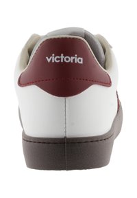 Sneaker bianco con accenti bordeaux sul tallone e sul lato. Presenta una suola marrone testurizzata, colletto imbottito e branding "victoria" in bianco.