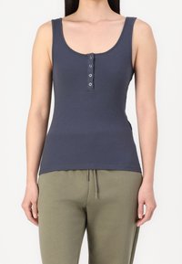 Kvinna i ärmlös marinblå ribbad topp med fyra knappar och olivgröna byxor med dragsko framför vit bakgrund.