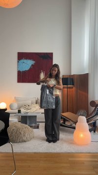 Indumento nero in confezione trasparente tenuto da una persona, circondato da un soggiorno moderno con un tavolo in marmo, texture morbide e illuminazione calda.