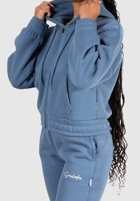 Sudadera con cremallera de color azul claro, con una gran capucha, bolsillos frontales y puños elásticos; combinada con pantalones a juego que presentan texto bordado.