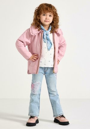 Ung pige med krøllet hår iført en pink jakke, hvid mønstret sweater, blåt tørklæde, jeans broderet med sommerfugle og pink støvler står selvsikkert.