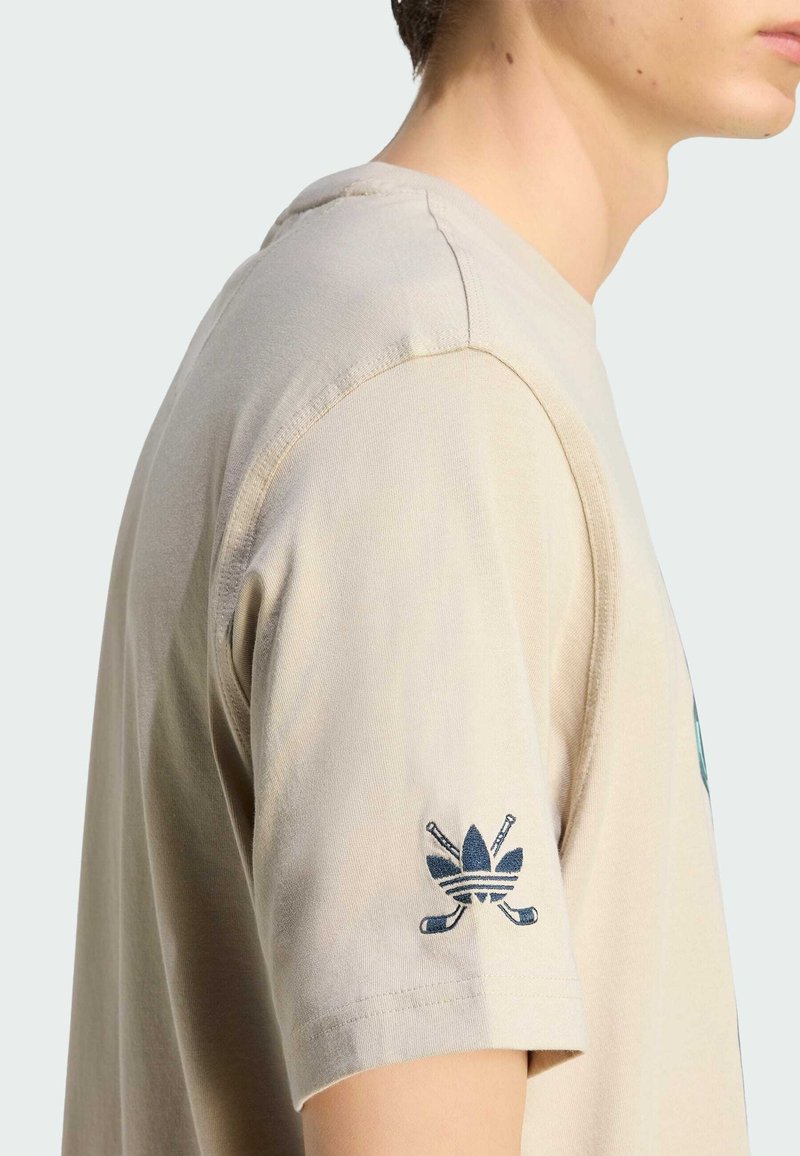 Maglietta beige in cotone con scollo rotondo, maniche corte e logo ricamato blu scuro sulla manica sinistra che rappresenta bastoni da golf incrociati.