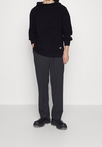 Hoodie noir en maille avec cordon de serrage, associé à un pantalon ample gris foncé et des bottes noires à semelle épaisse. Design simple avec des textures subtiles.