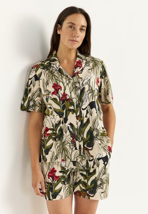 Vrouw draagt beige blouse met knopen en korte broek met print van groene bladeren, rode bloemen en zwart-witte vogels en dieren, staand tegen een muur.