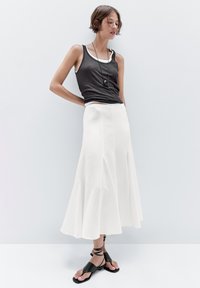 Jeune femme s'appuyant contre un mur blanc, portant un débardeur noir, une jupe midi blanche, des sandales noires et un long collier pendentif noir.