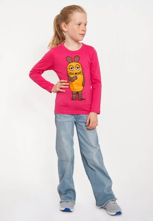 Junges Mädchen mit Pferdeschwanz, das ein leuchtend pinkfarbenes Langarmshirt mit einer Cartoon-Maus, helle blaue Jeans und graue Turnschuhe trägt und selbstbewusst dasteht.