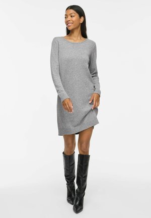 Femme souriante portant une robe tricotée grise à manches longues et des bottes noires montantes, posant devant un fond blanc uni.