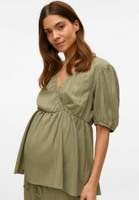 Top green per maternità con scollatura a V, vita arricciata e maniche a sbuffo. Il tessuto ha un aspetto leggero e texturizzato con sottili righe.