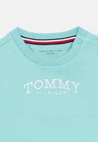 Heledu sinine puuvillane dressipluus, millel on soonikuga kaelus, tikitud "TOMMY HILFIGER" logo ja triibuline aktsent kaeluse sisepindadel.