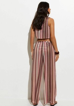 Vrouw met lang donker haar, gekleed in een mouwloos roze en bruin gestreept cropped topje en wijde broek, staand tegen een witte achtergrond.