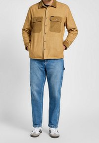 Bruine, gemêleerde flanellen shirtjas met twee borstzakken, gecombineerd met lichtblauwe rechte spijkerbroek en witte sneakers met zwarte strepen.