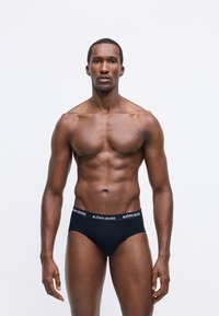 Svalnatý muž stojaci na bielom pozadí v čiernych björn borg slipsoch, stojaci čelom k objektívu s neutrálnym výrazom a uvoľnenými rukami pozdĺž tela.