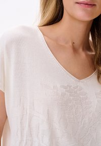 Helltaupe gestricktes Oberteil mit V-Ausschnitt und kurzen Ärmeln, das ein geprägtes Blumenmuster und eine weich strukturierte Oberfläche aufweist.