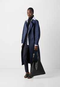 Long navy trench coat with double-breasted design, black turtleneck, striped shirt, dark trousers, and black tote bag. Smooth fabric and sleek silhouette.