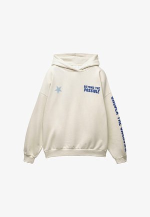 Kerman huppari, jossa on sininen tähti rinnassa, etupuolella oikealla teksti "MENNÄ YLI MAHDOLLISUUKSIEN" ja vasemmassa hihassa alaspäin menevä teksti "AVAA TUNNEMATON".