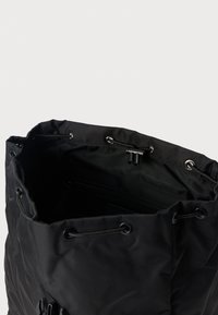 Μαύρη υφασμάτινη τσάντα με κλείσιμο με κορδόνι, διαθέτοντας υφή στην επιφάνεια και μεταλλικά κρικάκια για πρόσθετη λεπτομέρεια. Ορατή εσωτερική τσέπη.