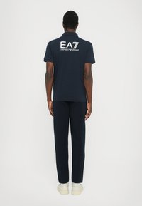 Polo navy de mangas curtas com o logótipo "EA7 Emporio Armani" em branco nas costas, combinado com calças navy coordenadas e ténis claros.